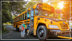 Children with backpacks boarding yellow school bus with safety arm activated, on treelined street with sun shining.